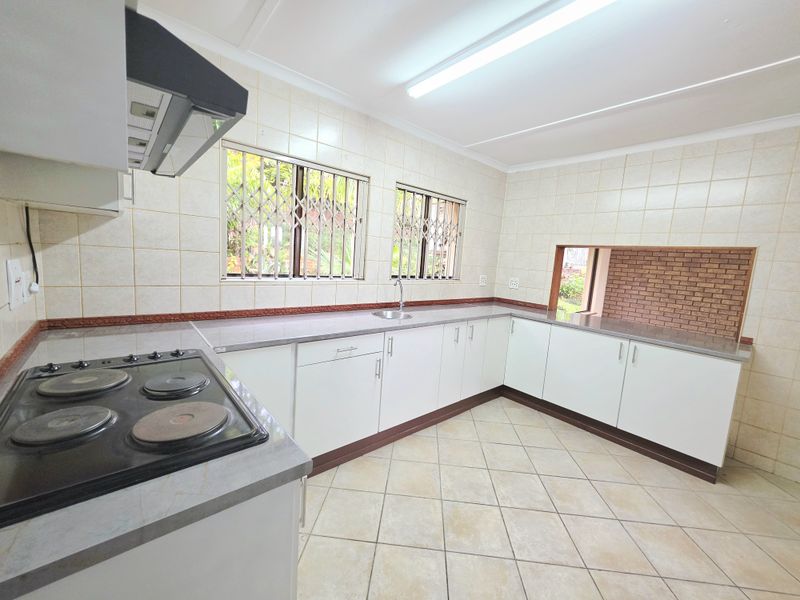 A functional kitchen featuring white cabinetry, an electric hob with extractor fan, tiled splashbacks, and a pass-through window to the adjacent living area.