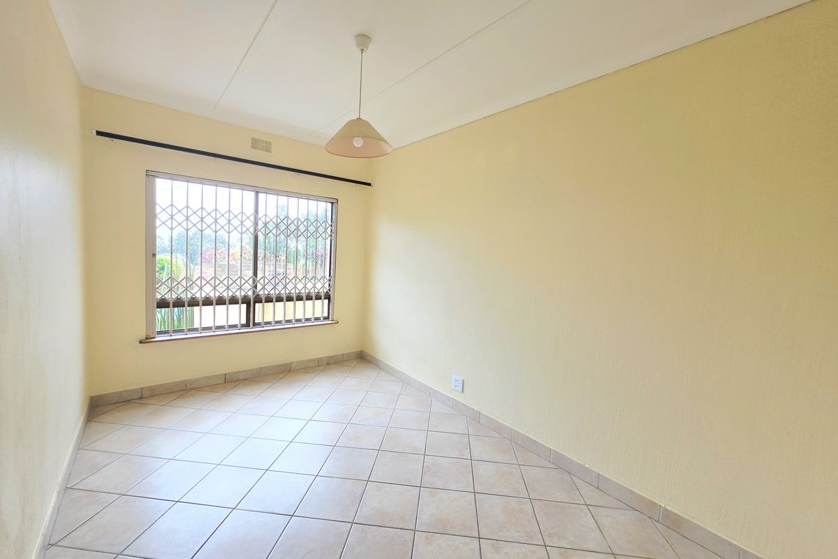 A bright, empty bedroom featuring neutral tiled flooring, a security gate on the window, and a simple pendant light fixture.