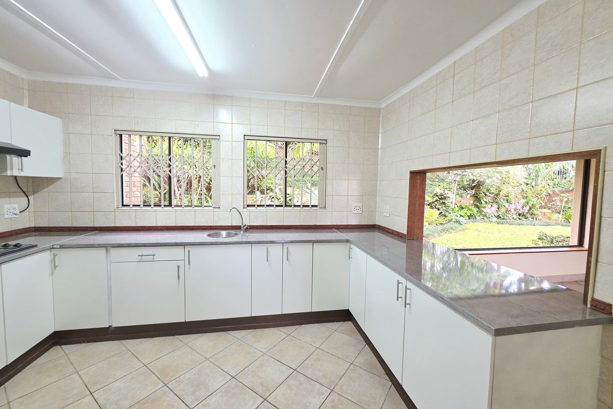 A functional kitchen featuring white cabinetry, laminate countertops, tiled surfaces, and a large window overlooking the garden, providing ample natural light and workspace.