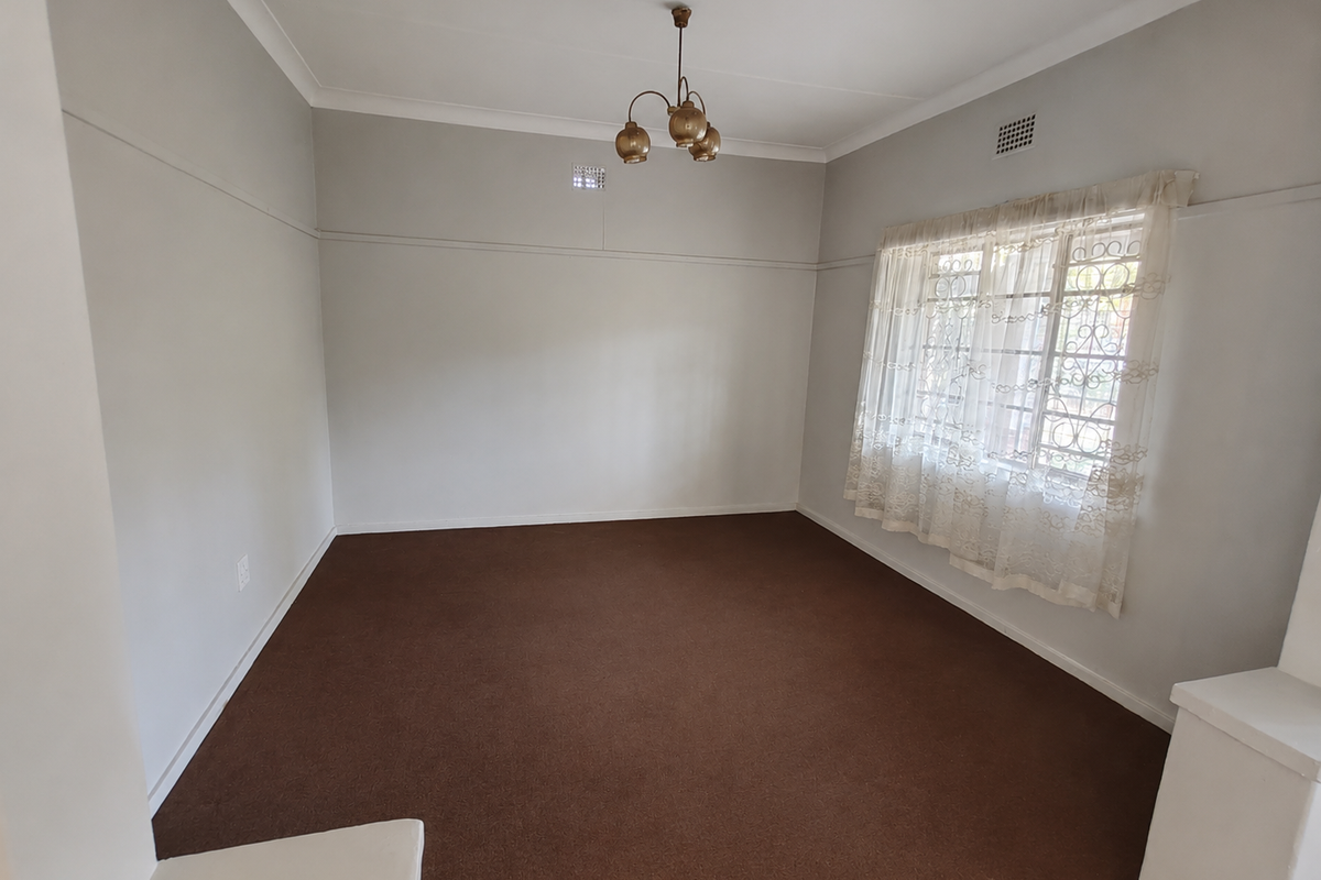 A spacious, empty living room featuring neutral walls, carpeted flooring, a classic ceiling light fixture, and a window fitted with a security gate.