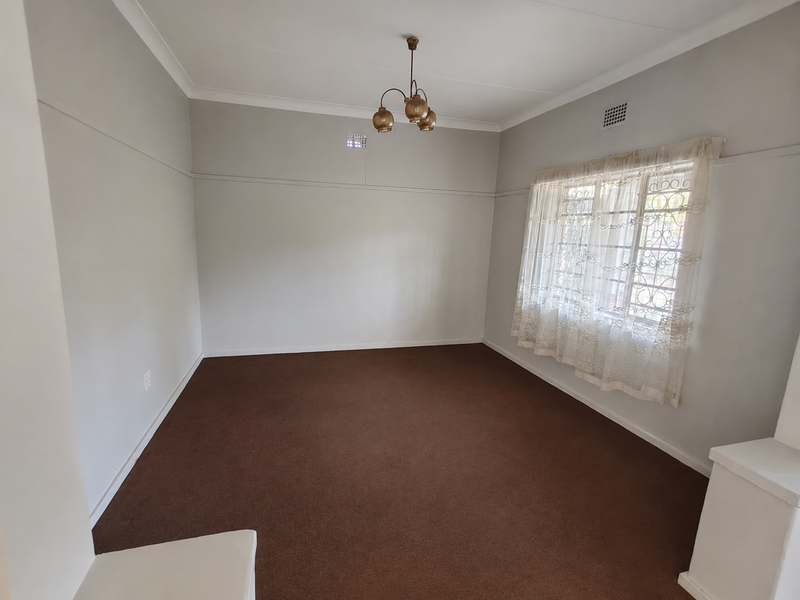 A spacious, empty living room featuring neutral walls, carpeted flooring, a classic ceiling light fixture, and a window fitted with a security gate.