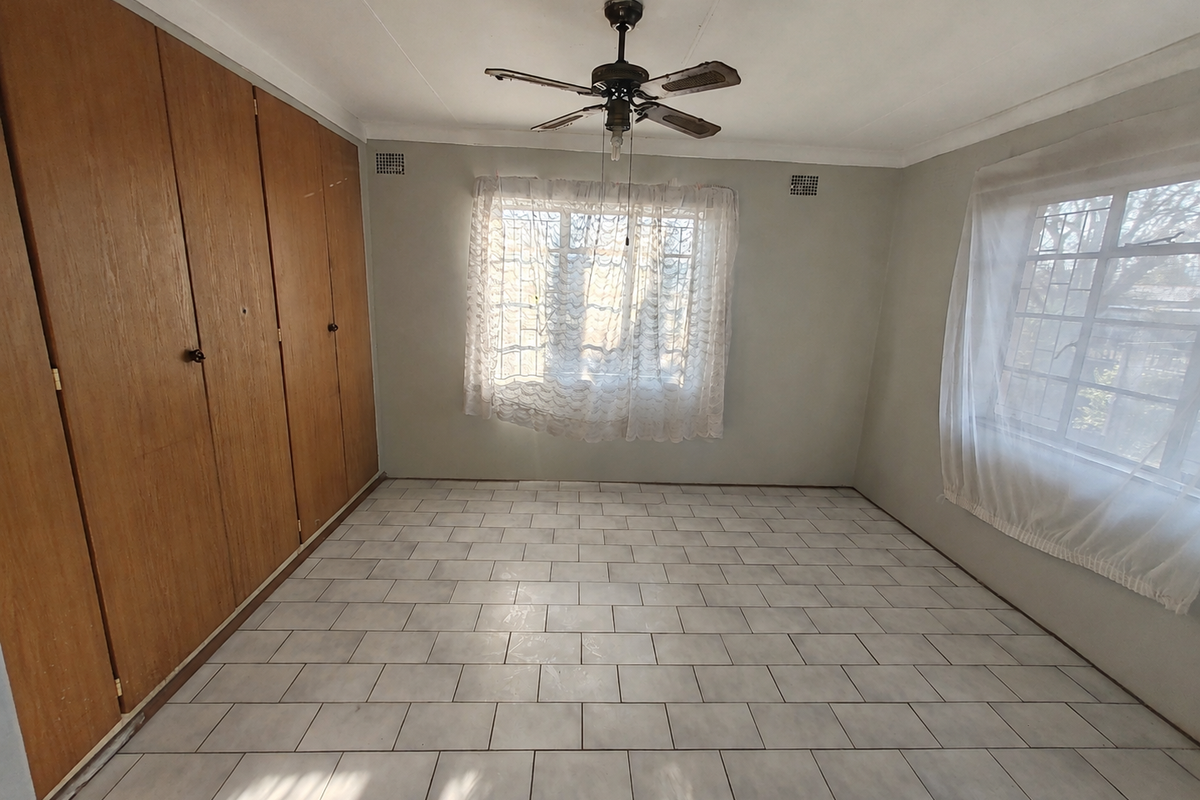 An empty bedroom featuring tiled flooring, built-in wooden cupboards, a ceiling fan, and windows fitted with security bars.