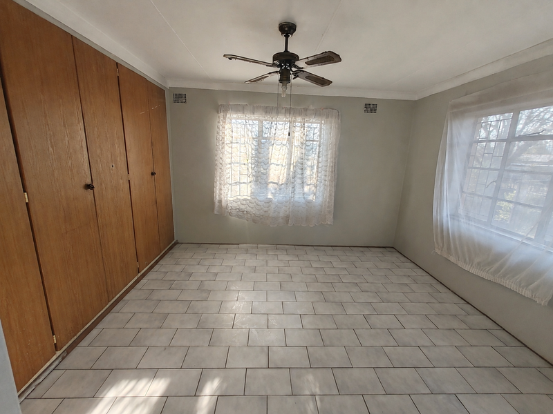 An empty bedroom featuring tiled flooring, built-in wooden cupboards, a ceiling fan, and windows fitted with security bars.