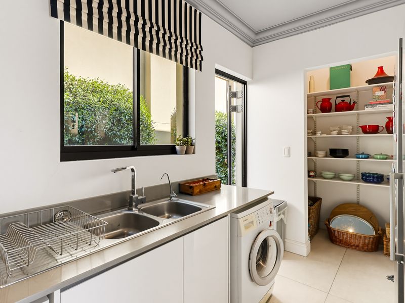 A functional scullery featuring a stainless steel double sink, built-in cabinetry, laundry plumbing, and an integrated walk-in pantry with ample shelving.