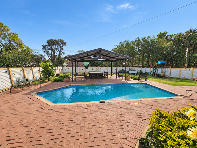 Spacious outdoor entertainment area featuring a swimming pool, a large covered patio structure, and a well-maintained paved terrace surrounded by garden fencing.