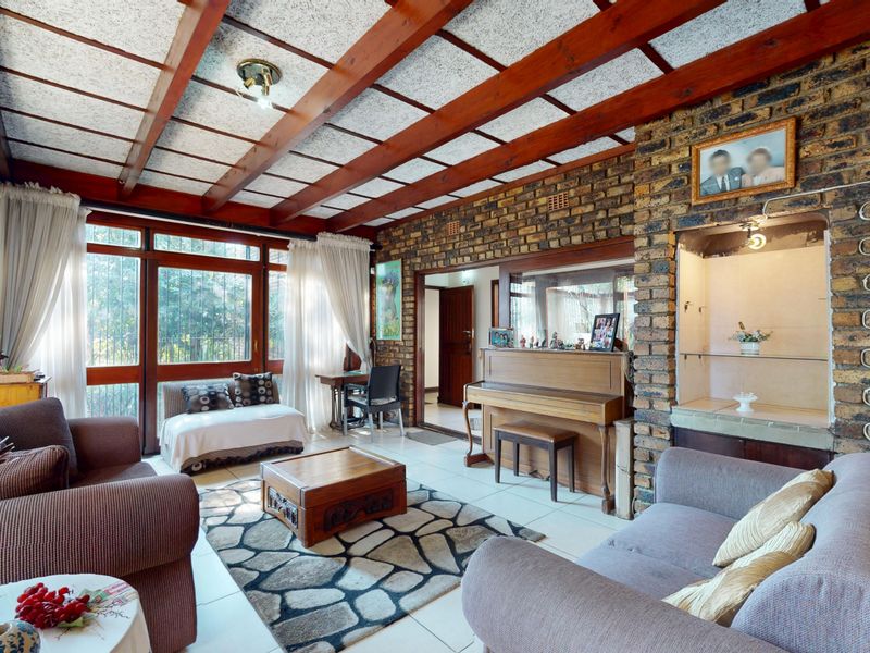A spacious living room featuring exposed timber ceiling beams, a rustic brick feature wall with a built-in display niche, and large windows.