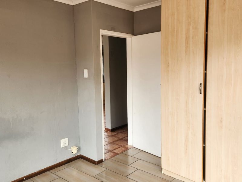 An empty bedroom featuring tiled flooring, a built-in wooden wardrobe, and neutral grey walls with access to an adjoining hallway.