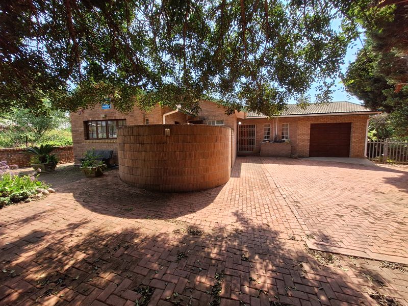 Front exterior view of a brick house featuring a paved driveway, integrated garage, and a curved brick feature wall under mature tree shade.