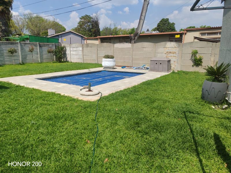 A backyard area featuring a swimming pool with a cover, surrounded by a lawn and a precast concrete boundary wall.