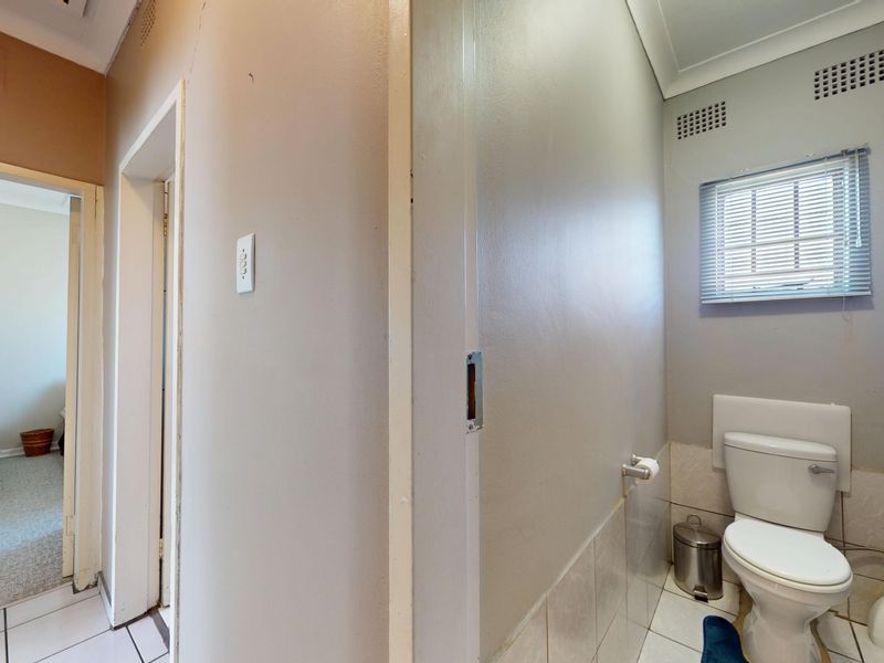 A functional bathroom featuring a standard toilet, tiled flooring, and a window with blinds, adjacent to a hallway leading to a bedroom.