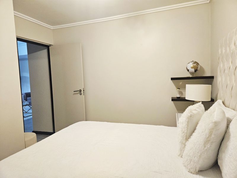 A well-maintained bedroom featuring neutral wall tones, crown molding, and built-in floating shelves, with a doorway leading to an adjacent living space.