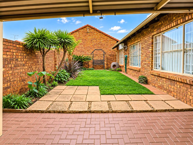 A well-maintained rear courtyard featuring a paved patio area, a neat lawn, and brick boundary walls, viewed from under a covered patio.