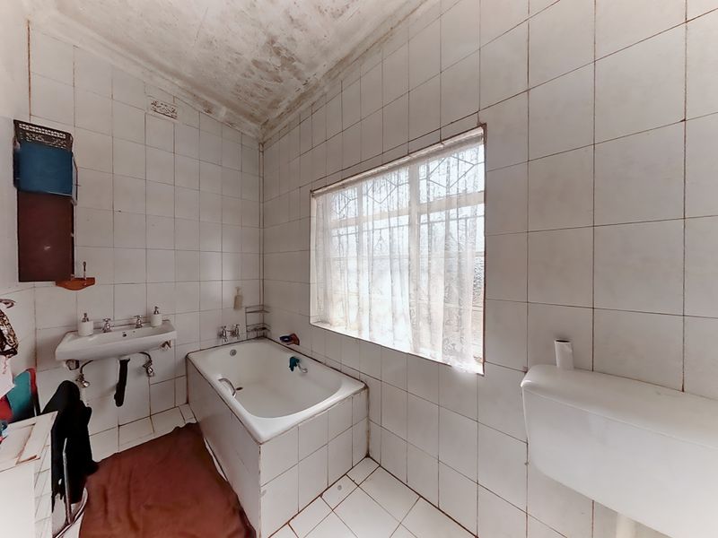 A dated bathroom featuring a built-in bathtub, wall-mounted basin, and full-height wall tiling, requiring renovation due to visible ceiling damage and worn finishes.