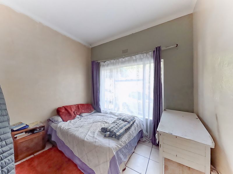A bedroom featuring tiled flooring and a large window with a curtain rail, currently showing signs of wear on the wall finishes.