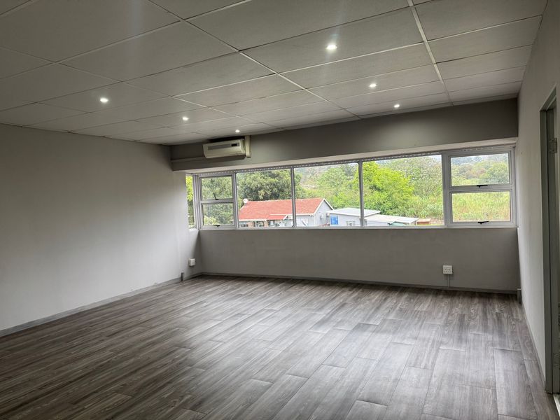 A bright, empty living area featuring modern laminate wood flooring, a drop ceiling with recessed lighting, and a large window with exterior views.