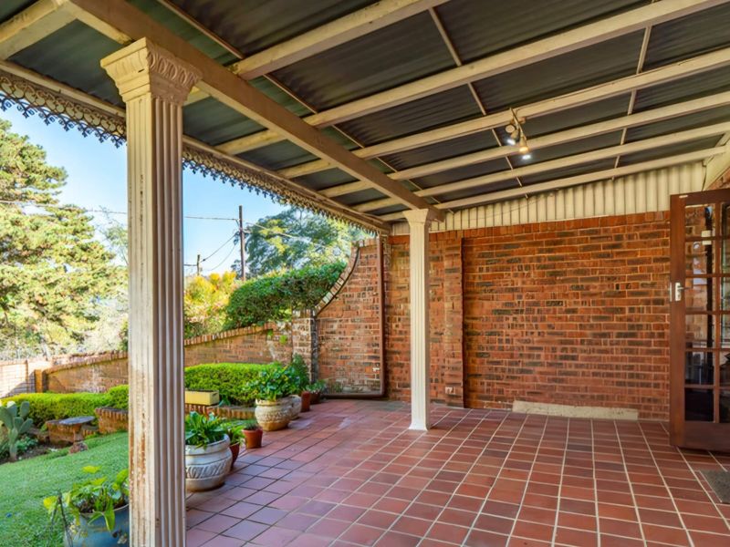 A covered outdoor patio featuring brick walls, tiled flooring, and decorative support columns, overlooking a landscaped garden area.