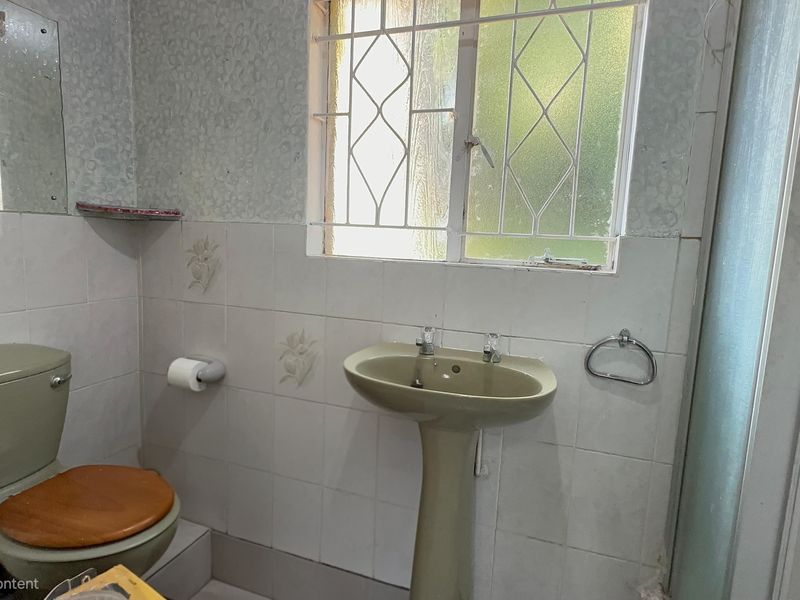 A dated bathroom featuring a pedestal basin, toilet, and shower enclosure with tiled walls and security bars on the window.