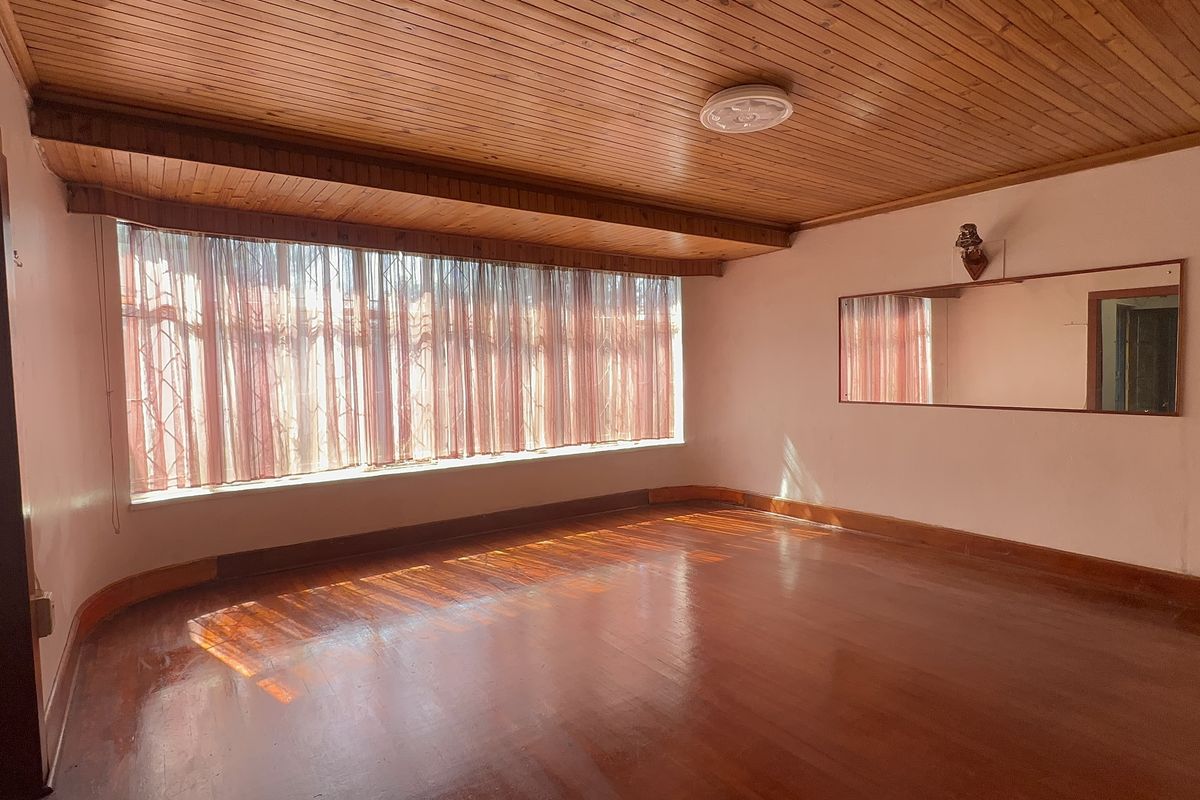 A spacious, empty living room featuring polished hardwood flooring, a timber-paneled ceiling, and a large window with security bars.