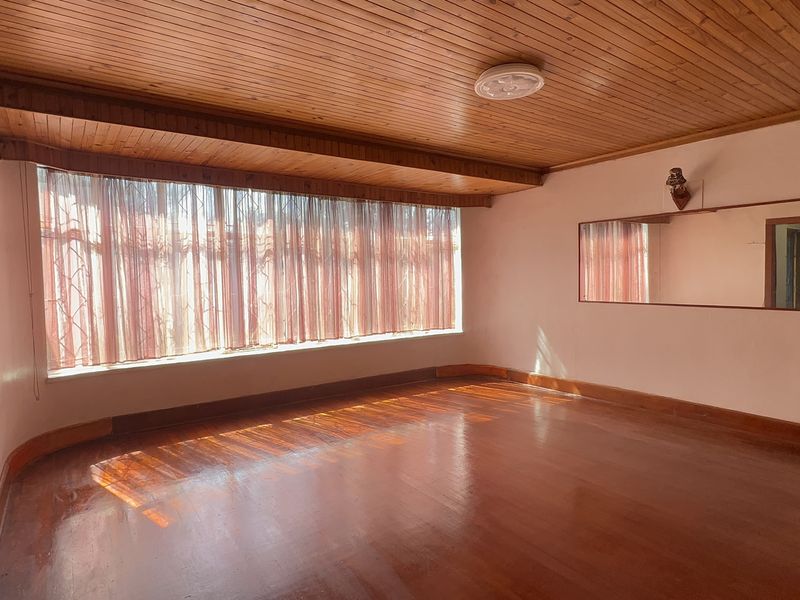 A spacious, empty living room featuring polished hardwood flooring, a timber-paneled ceiling, and a large window with security bars.
