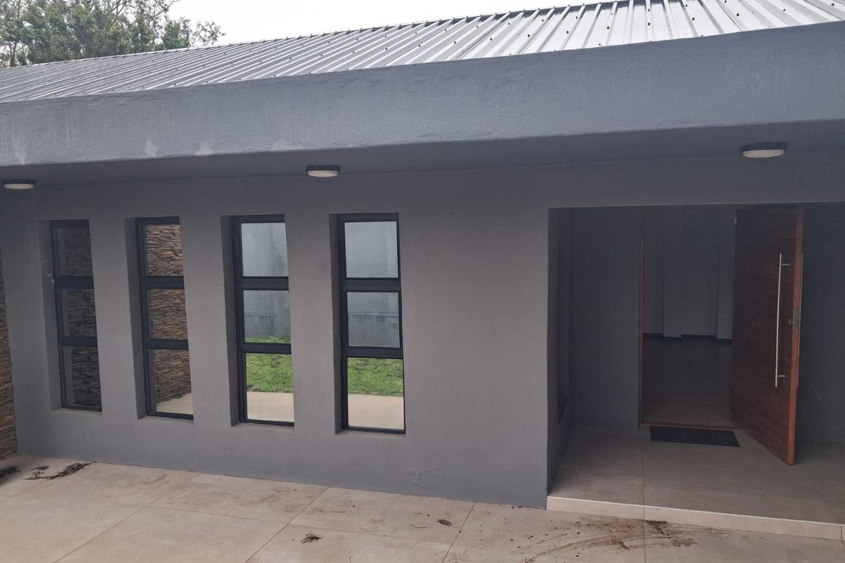 A modern rear exterior view featuring a grey rendered facade, metal roof, aluminium windows, and a timber entrance door leading to a tiled patio area.