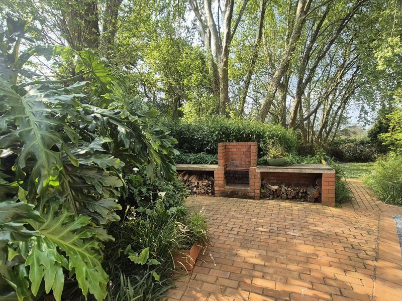 A tranquil garden area featuring a brick-built outdoor braai and a paved patio surrounded by mature trees and lush greenery.