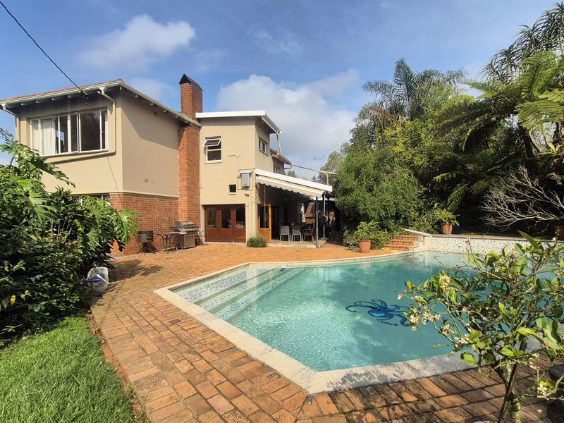 Rear view of the house featuring a swimming pool, brick paved patio area, and a retractable awning, set within a landscaped garden.