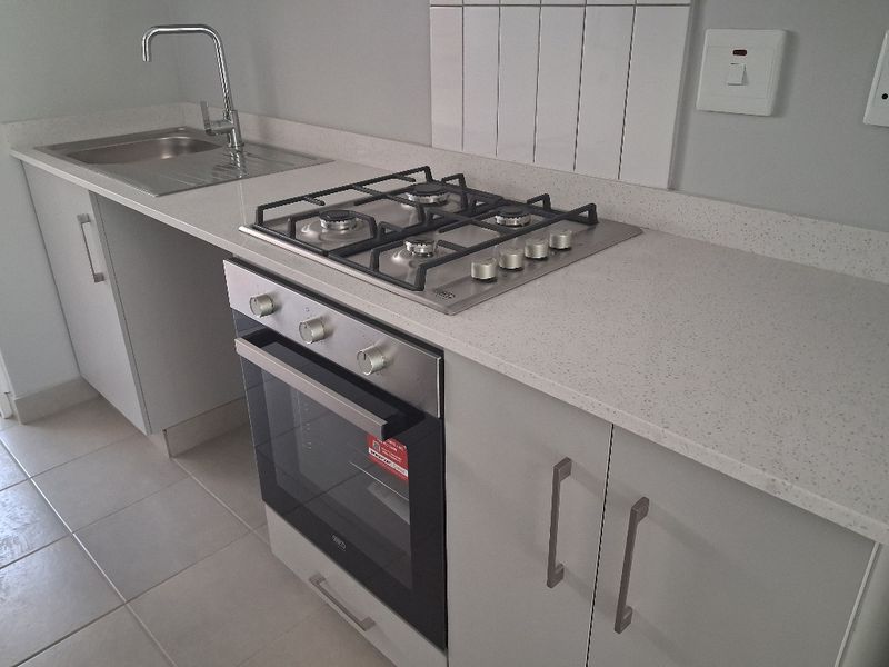 A modern, clean kitchen area featuring a stainless steel sink, gas hob, built-in oven, and stone countertops with neutral cabinetry.