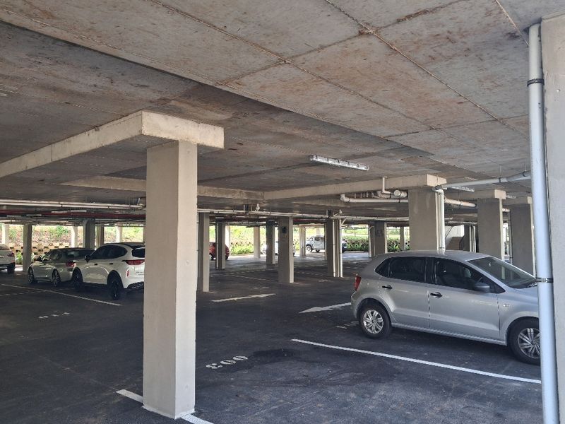 A covered basement parking garage featuring concrete structural elements, exposed utility piping, and marked parking bays.
