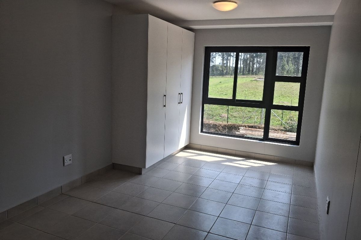 A bright, empty bedroom featuring neutral tiled flooring, a built-in wardrobe, and a large window with views of the surrounding greenery.
