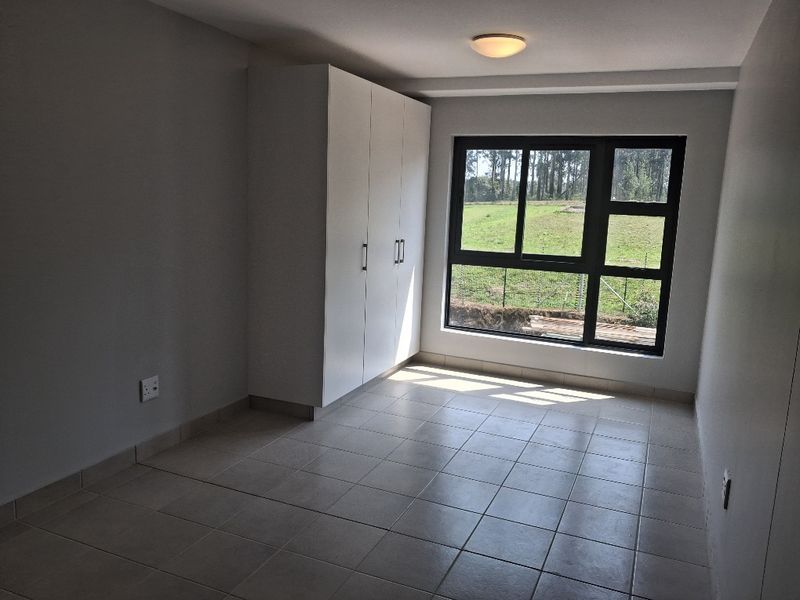 A bright, empty bedroom featuring neutral tiled flooring, a built-in wardrobe, and a large window with views of the surrounding greenery.