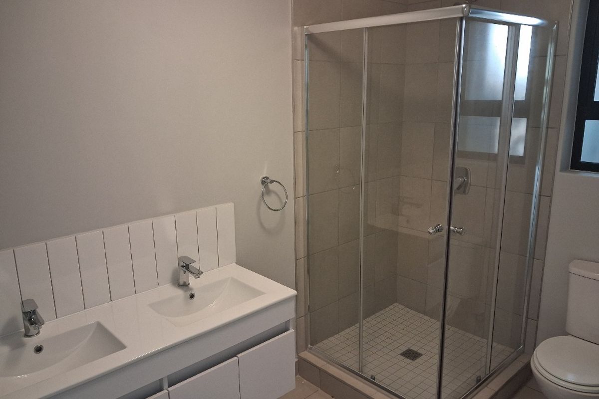 Modern bathroom featuring a double vanity with white cabinetry, a glass-enclosed shower, and a standard toilet with neutral tiled finishes.