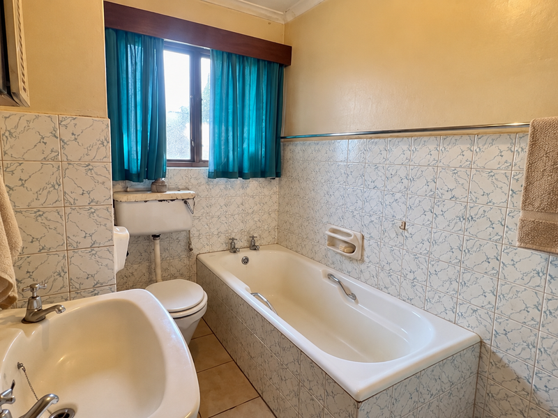 A dated bathroom featuring a standard bathtub, pedestal sink, and toilet with original patterned wall tiling and neutral floor tiles.