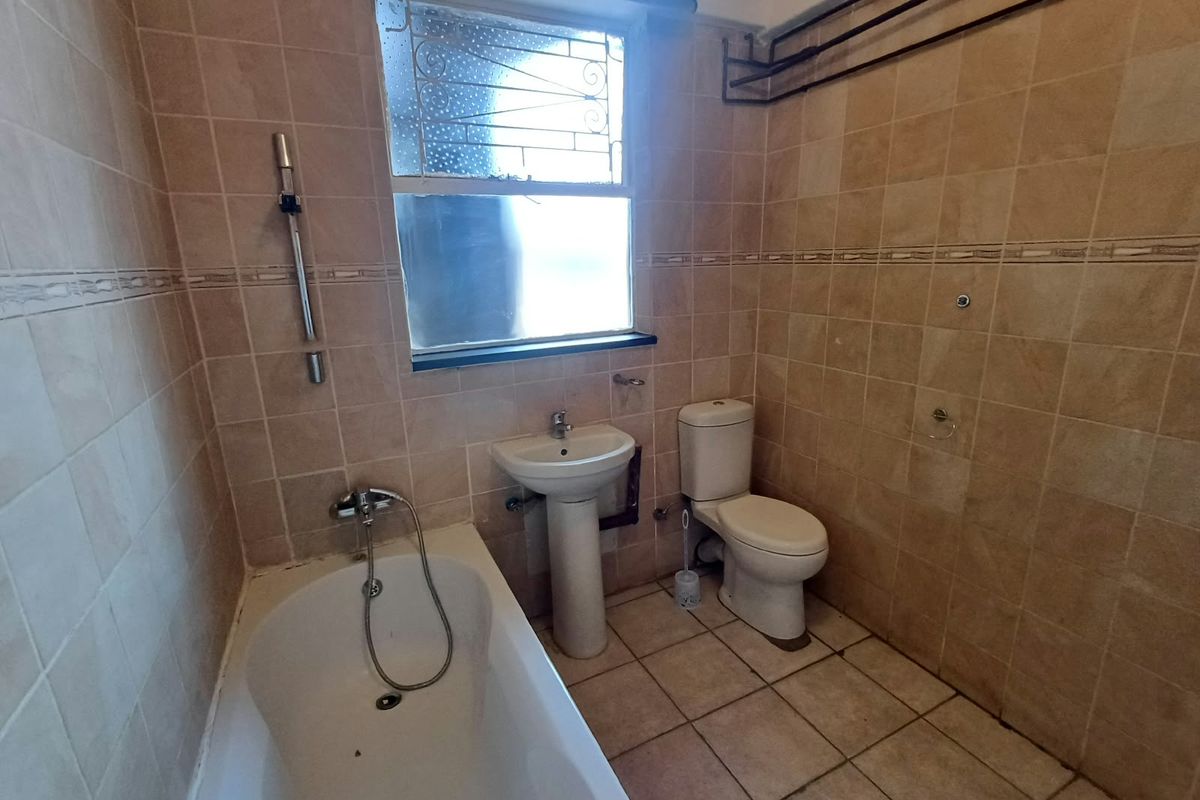 A dated bathroom featuring a pedestal basin, toilet, and bathtub with full-height ceramic wall tiling and a window with security bars.