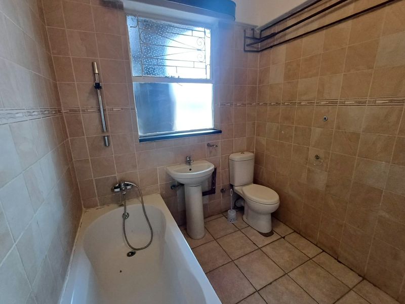 A dated bathroom featuring a pedestal basin, toilet, and bathtub with full-height ceramic wall tiling and a window with security bars.