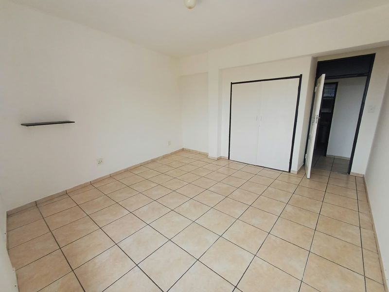 An empty bedroom featuring neutral tiled flooring, a built-in wardrobe, and a wall-mounted shelf, offering a clean and versatile space.