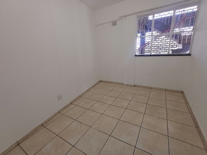 An empty bedroom featuring neutral tiled flooring, white walls, and a window fitted with security bars.