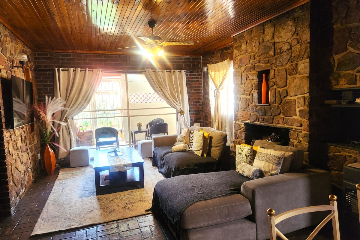 Cozy living room featuring a rustic stone fireplace, exposed brick walls, warm timber ceiling, and terracotta tile flooring with access to an outdoor area.