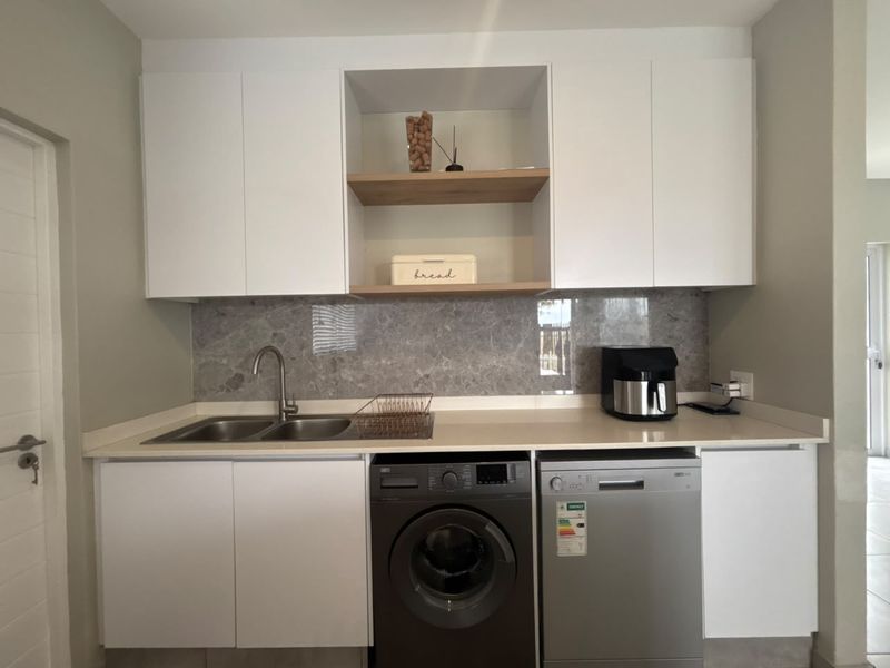 A modern scullery featuring white cabinetry, stone countertops, a stainless steel sink, and a stone splashback, providing efficient utility and prep space.