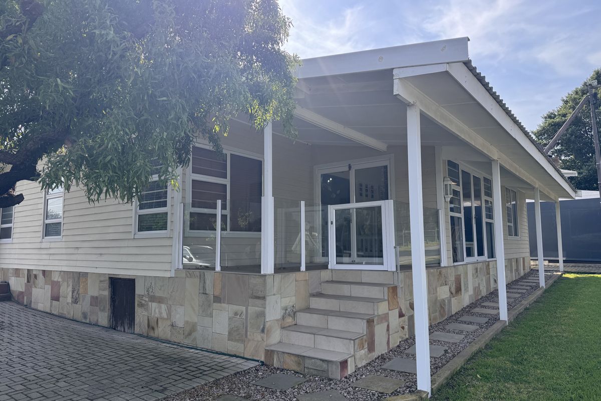 A well-maintained rear exterior featuring a covered patio with glass balustrades, stone-clad base, and paved access, set against a tidy garden.