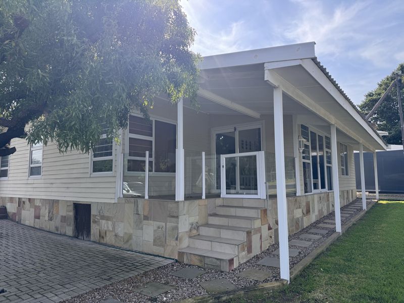 A well-maintained rear exterior featuring a covered patio with glass balustrades, stone-clad base, and paved access, set against a tidy garden.