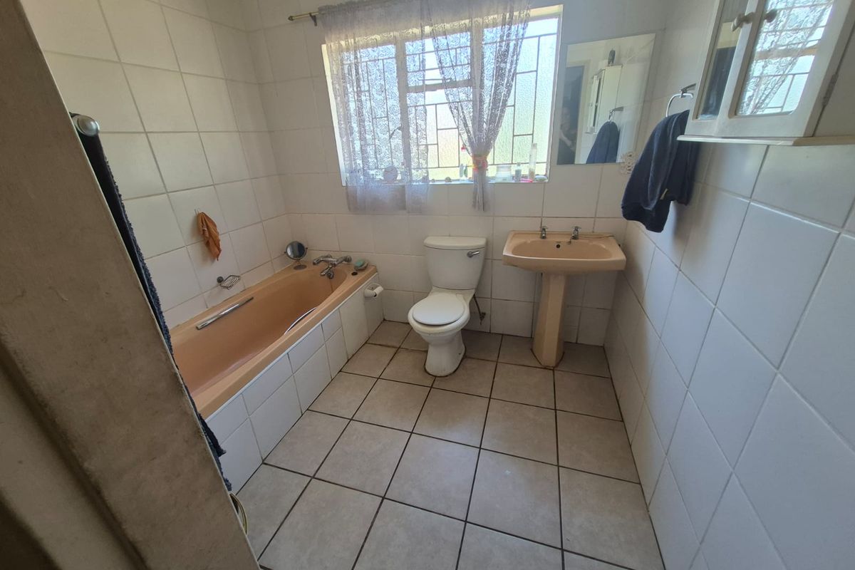 A dated bathroom featuring a peach-colored bathtub and pedestal sink, tiled walls and flooring, and a window with security bars.