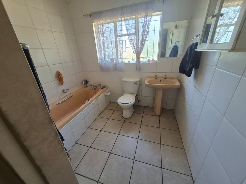 A dated bathroom featuring a peach-colored bathtub and pedestal sink, tiled walls and flooring, and a window with security bars.