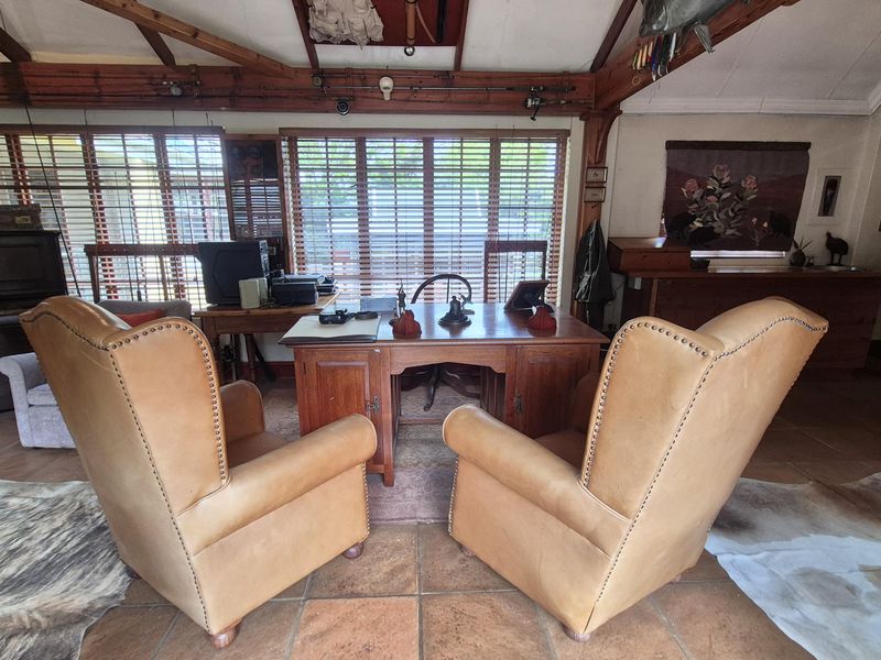 A rustic home office featuring exposed timber ceiling beams, terracotta floor tiles, and large windows with venetian blinds.