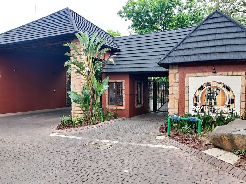 The entrance to Savuti Sands apartment complex featuring a paved driveway, security gate, and a well-maintained guard house with stone and tile finishes.