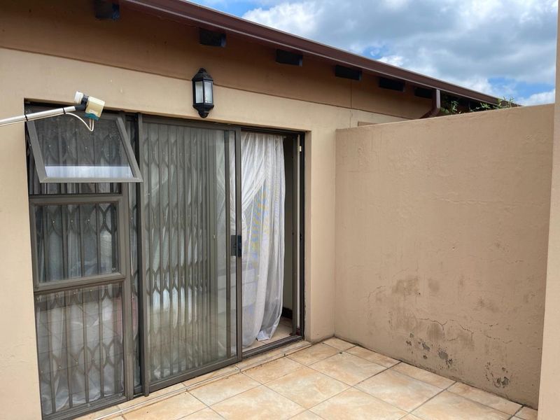 A small paved patio area featuring a sliding glass door with security gate and exterior wall lighting, showing signs of wear on the wall finish.