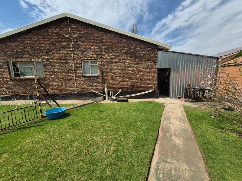 Rear view of the property featuring a face brick exterior, a corrugated metal lean-to structure, and a concrete pathway leading through the garden area.