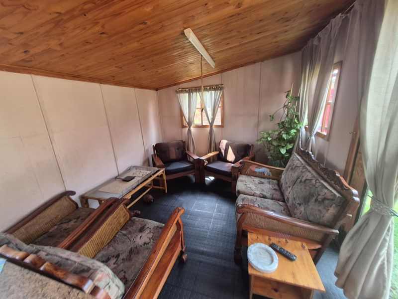 A living room featuring a timber-clad ceiling, tiled flooring, and natural light through casement windows, offering a rustic and dated aesthetic.