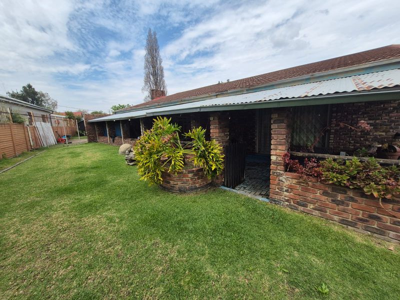 Rear view of a brick house featuring a covered patio area, brick pillars, and a spacious lawn with established garden beds.