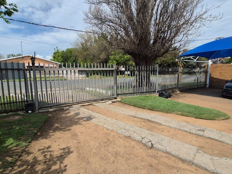 Front exterior view featuring a metal security gate, concrete driveway tracks, and a shade sail structure over the parking area.
