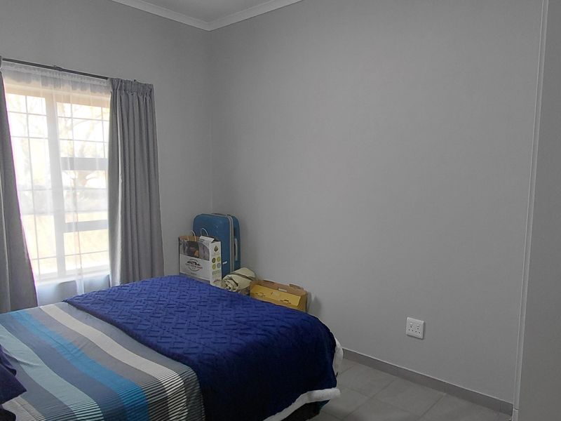 A simple bedroom featuring tiled flooring, neutral grey walls, and a window fitted with security bars.